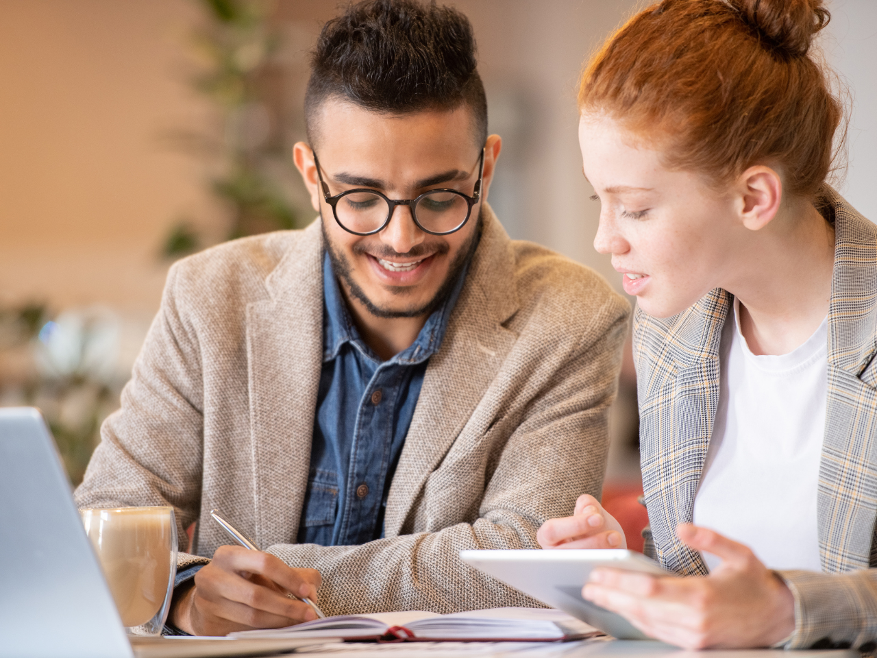 A young man and woman in business attire are collaborating at a table in a well-lit, modern workspace. The man, wearing glasses and a beige blazer over a denim shirt, is smiling while taking notes in a notebook. The woman, dressed in a plaid blazer, is holding a tablet and discussing something with him. A laptop and a coffee cup are also on the table, indicating a productive work session.